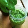 Een glazen potje vol veganistische pesto op een houten tafel met een basilicumplantje ernaast.